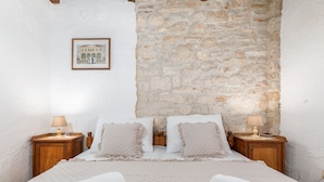 Quarto com parede de pedra, cama com roupa de cama branca, duas mesas de cabeceira com abajures e um quadro na parede.