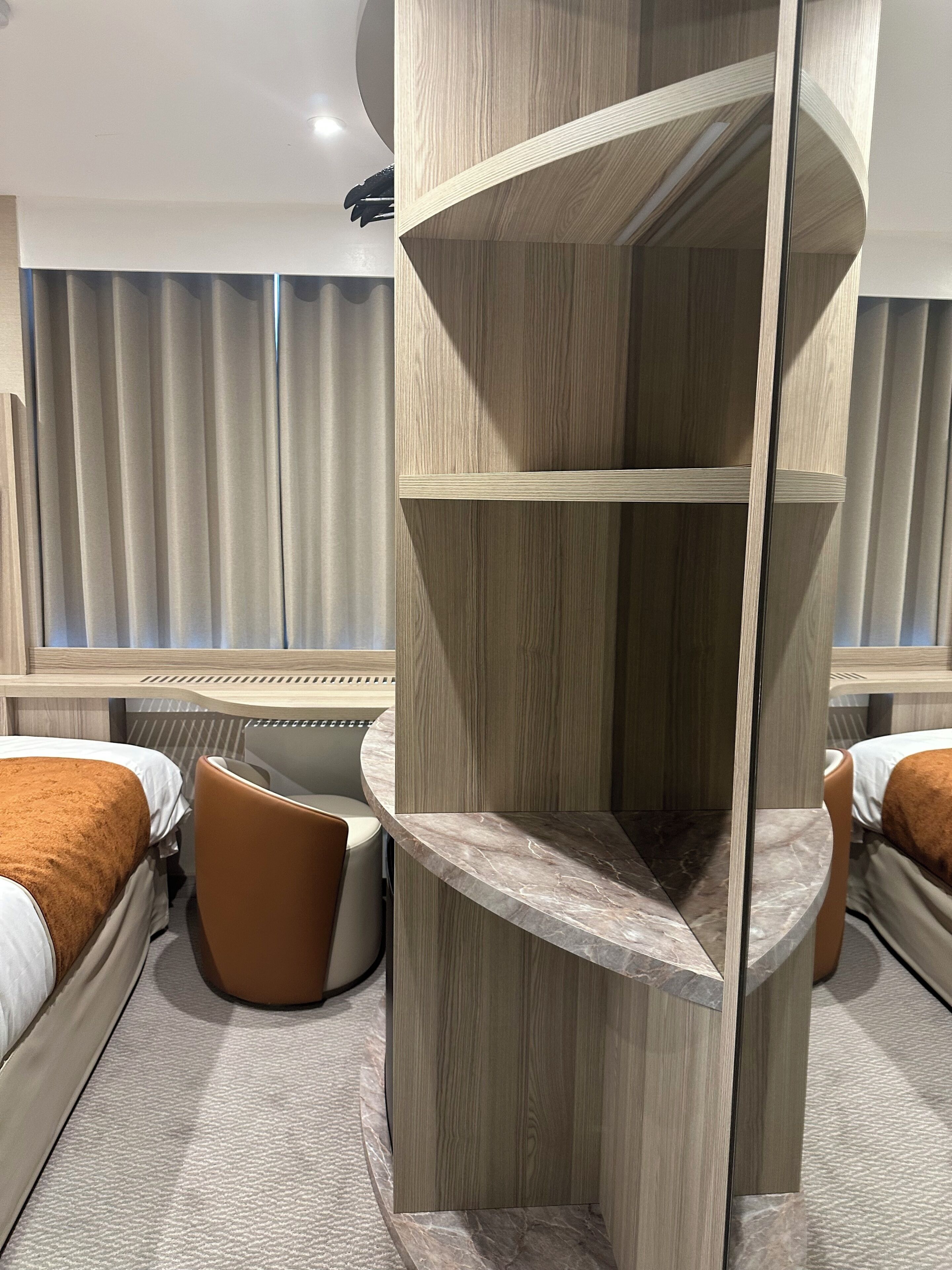 A modern hotel room with a curved wooden bed, a small seating area, and a large window with curtains.