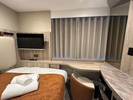 Ein Hotelzimmer mit Bett, Schreibtisch, Stuhl, Fernseher und einem Fenster mit Vorhängen.
