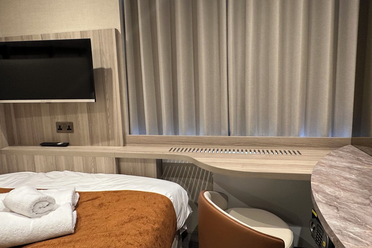 A hotel room with a bed, a TV mounted on the wall, a bathroom with a shower, and a desk with a chair.