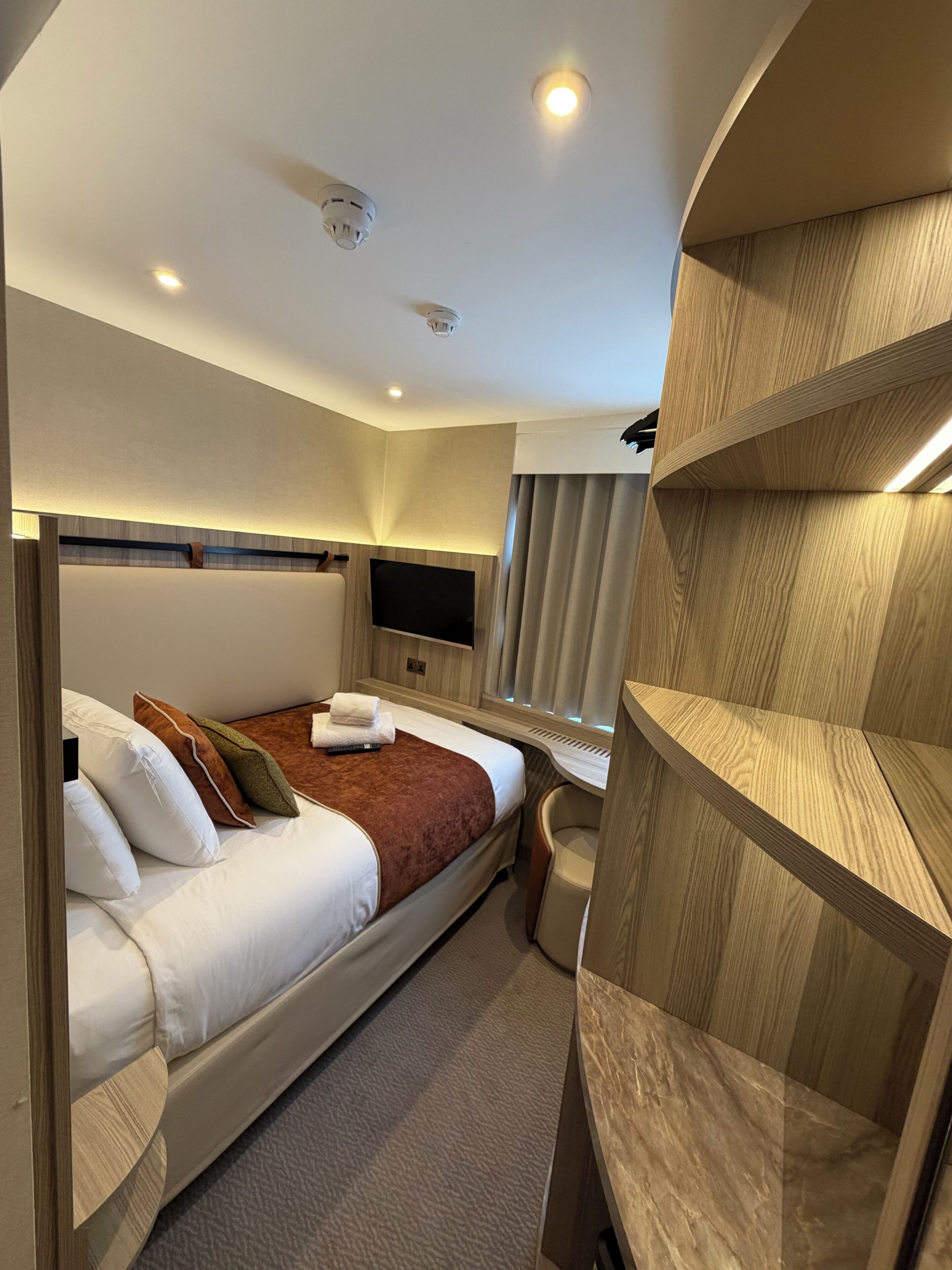 A modern hotel room with a bed, a desk, a chair, a television, and built-in shelving.