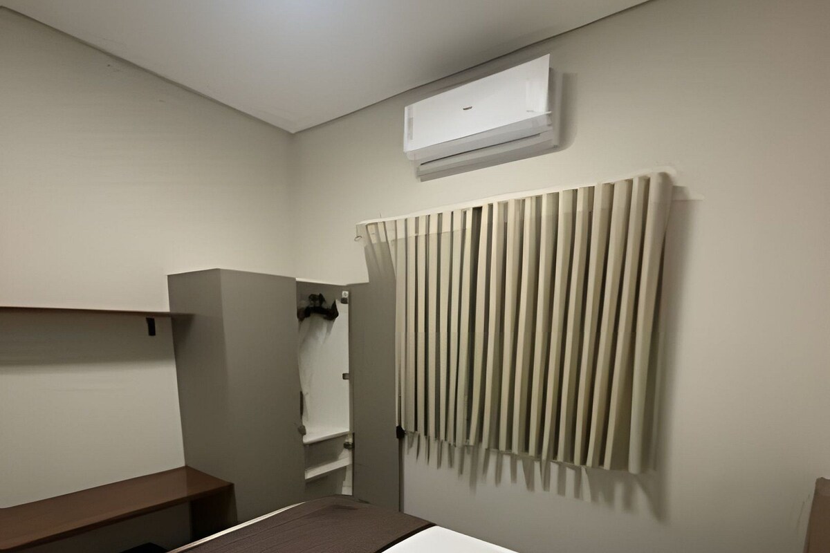 A modern hotel room with a bed, a built-in bench, a wall-mounted air conditioner, and a curtain.