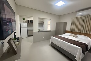A hotel room with a bed, a desk with a computer, a refrigerator, a microwave, and a window with blinds. - Studios Jocatita (Cuiabá)