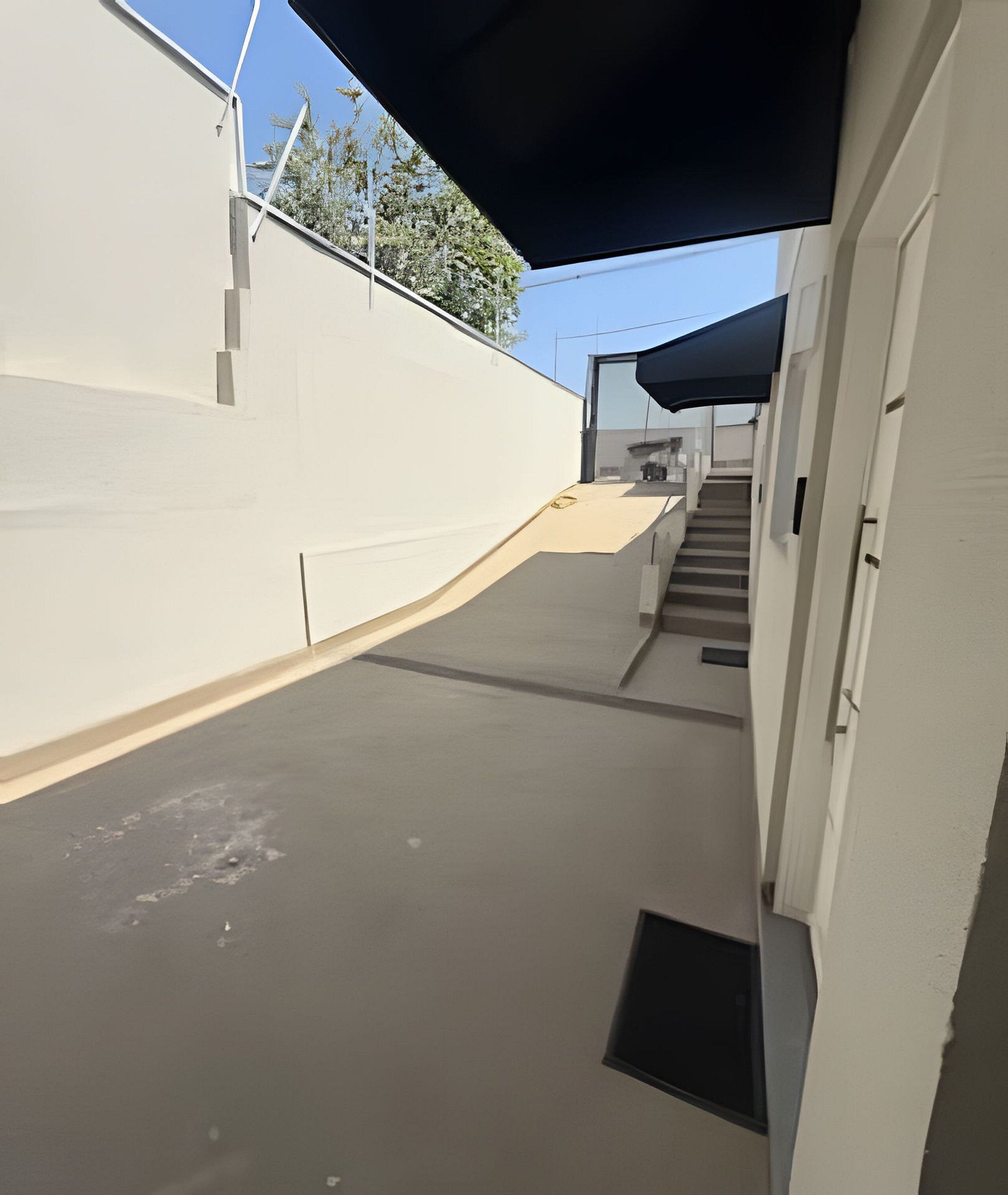 Um corredor exterior estreito com uma rampa que leva a uma entrada de edifício, rodeado por paredes brancas e um céu limpo.