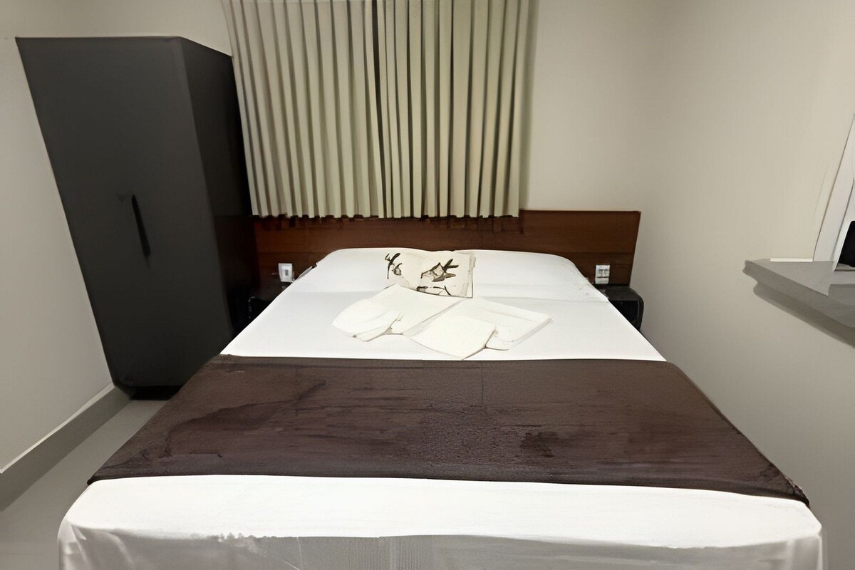 A hotel room with a bed, a wardrobe, a bedside table, and an air conditioning unit.