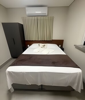 A hotel room with a bed, a wardrobe, a bedside table, and an air conditioning unit.