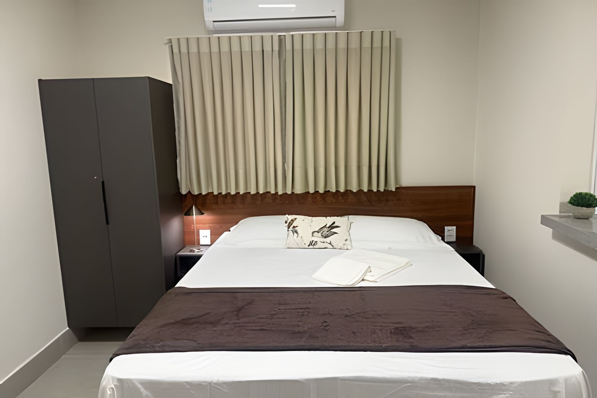 Quarto de hotel com cama, mesa de cabeceira, guarda-roupa e janela com cortinas.