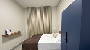 Una habitación de hotel con una cama, un armario azul, un estante con un diploma enmarcado y una cortina.