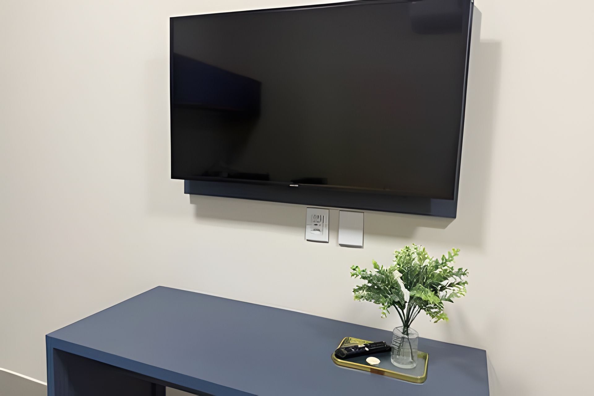 Un televisor de pantalla plana montado en la pared sobre un aparador azul oscuro con una planta pequeña y un control remoto.