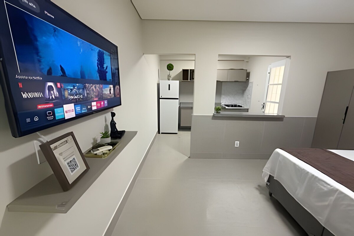 A modern hotel room with a flat-screen TV mounted on the wall, a bed, a small kitchenette, and a bathroom visible through an archway.