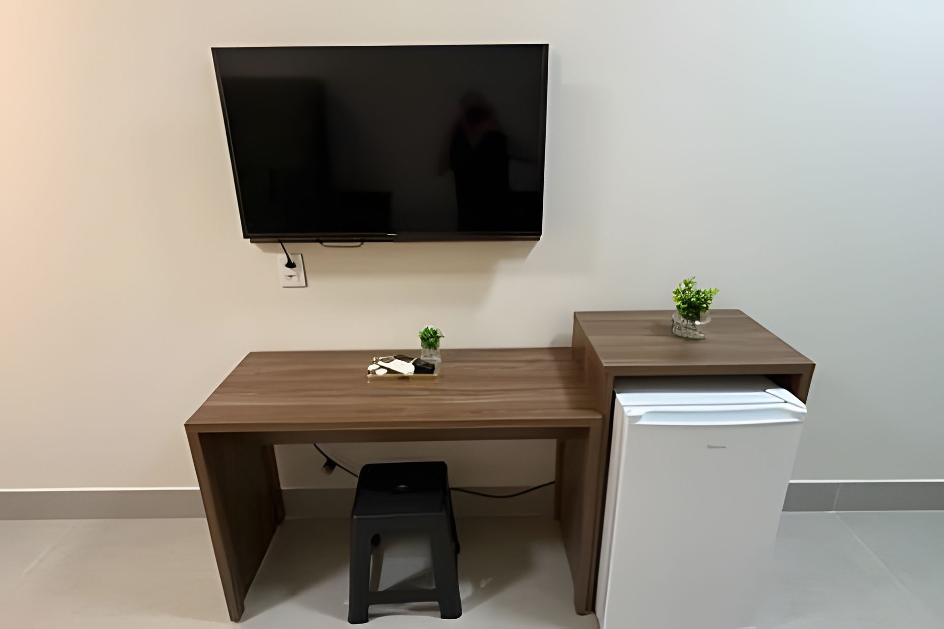 A compact workspace with a flat-screen TV mounted on the wall, a small desk with a plant, and a white mini-fridge.