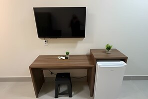 A compact workspace with a flat-screen TV mounted on the wall, a small desk with a plant, and a white mini-fridge.
