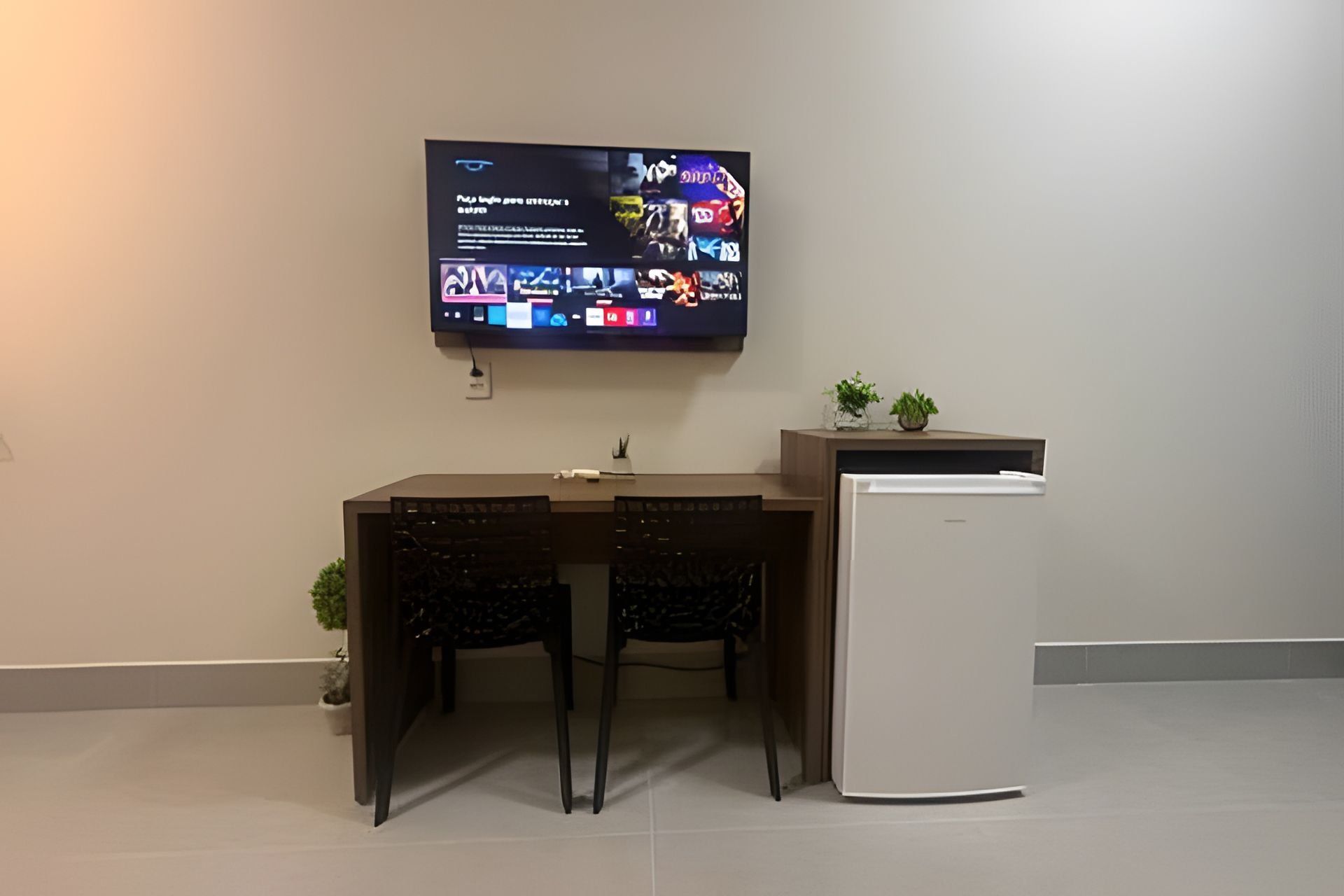 Eine moderne Küche mit Flachbildfernseher über einem dunklen Holztisch und Stühlen. Ein weißer Kühlschrank steht rechts, und auf der Arbeitsplatte befinden sich zwei kleine Topfpflanzen.