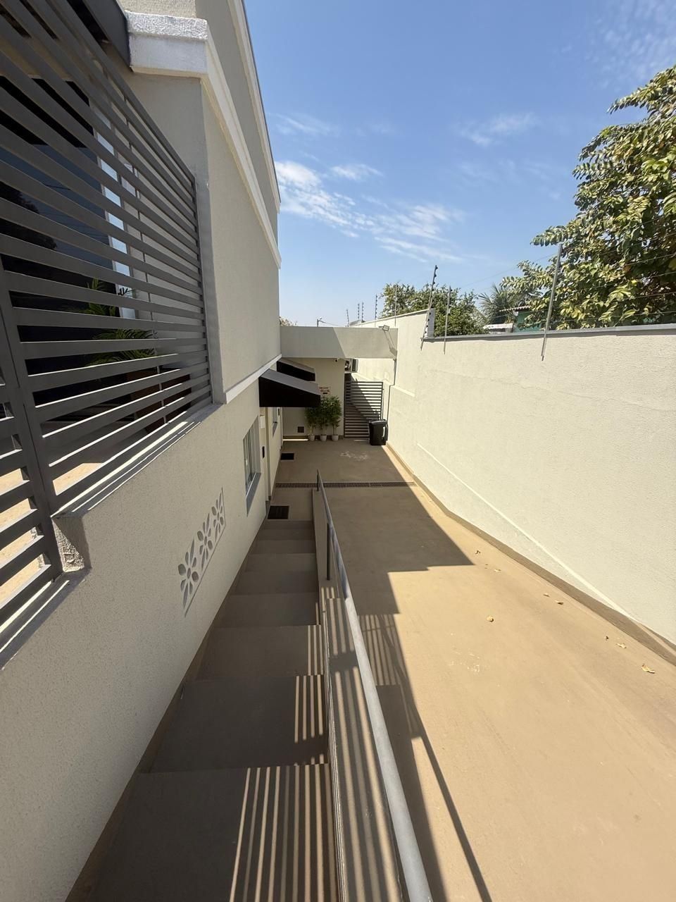 Um corredor externo estreito, com guarda-corrimão de metal, piso de concreto e parede branca com um logotipo.