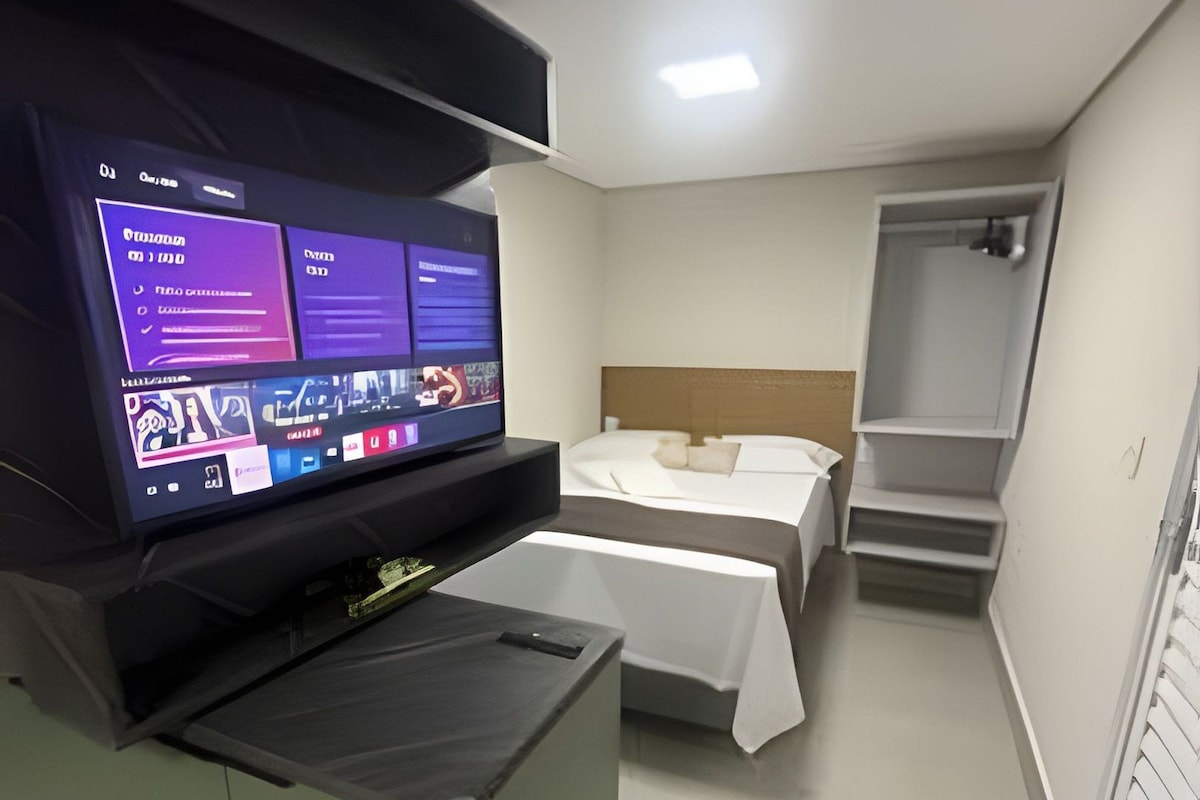 A hotel room with a bed, a desk with a computer, a TV, and a shower area.