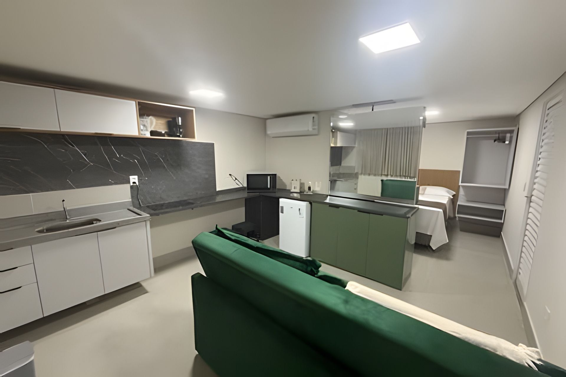 A compact kitchen with a refrigerator, microwave, and sink. There is a green sofa and a small dining area with a table and chairs.