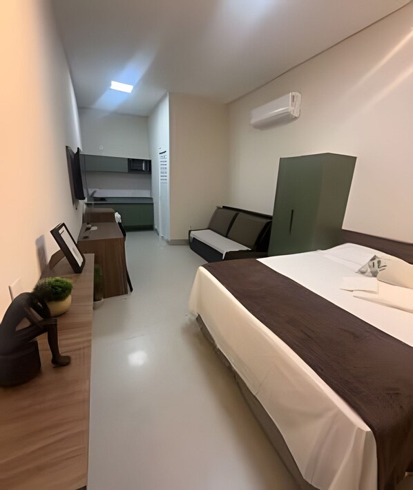 A compact hotel room with a bed, a desk, a chair, a small potted plant, a TV, and a kitchenette.