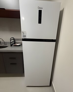 Ein weißer Kühlschrank mit schwarzer Bedienblende und einem Logo, der sich in einer Küche mit grauer Arbeitsplatte und zwei Schubladen befindet.