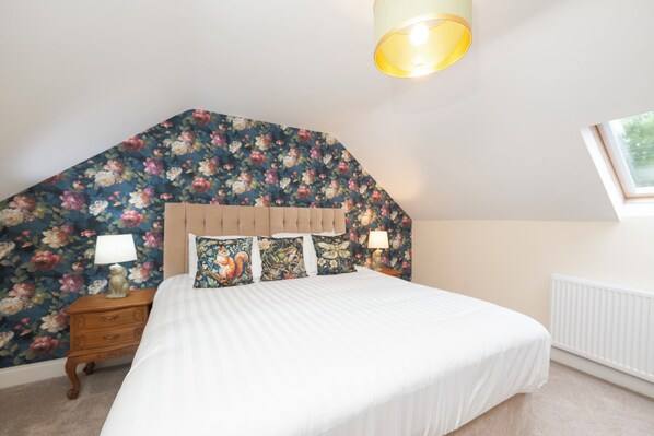 A bedroom with a sloped ceiling, floral wallpaper, a bed with white bedding, two bedside lamps, and a nightstand with a lamp.