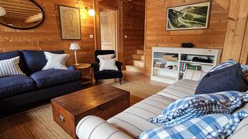 A cozy log cabin interior with a blue sofa, a striped armchair, a wooden coffee table, and a bookshelf.