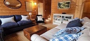 A cozy log cabin interior with a blue sofa, a striped armchair, a wooden coffee table, and a bookshelf.