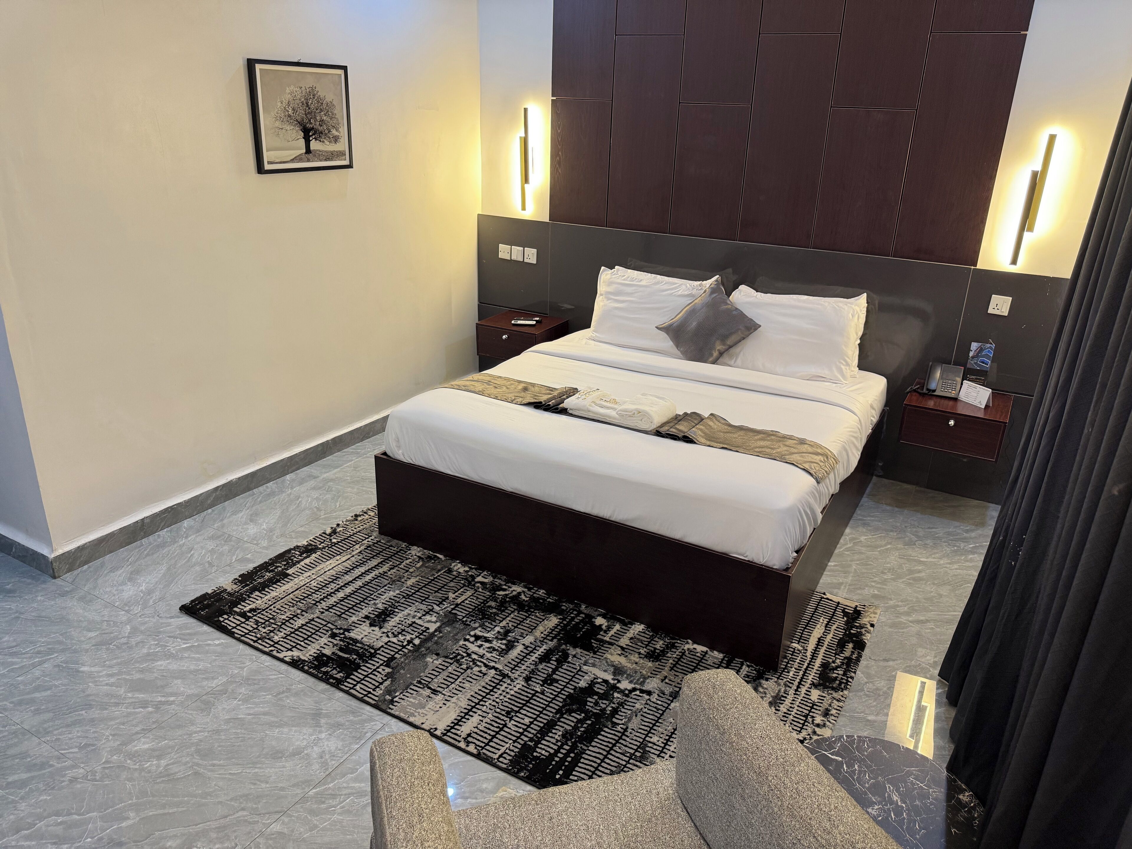 A modern hotel room with a large bed, two bedside tables, a nightstand, a wall-mounted light, and a framed picture.
