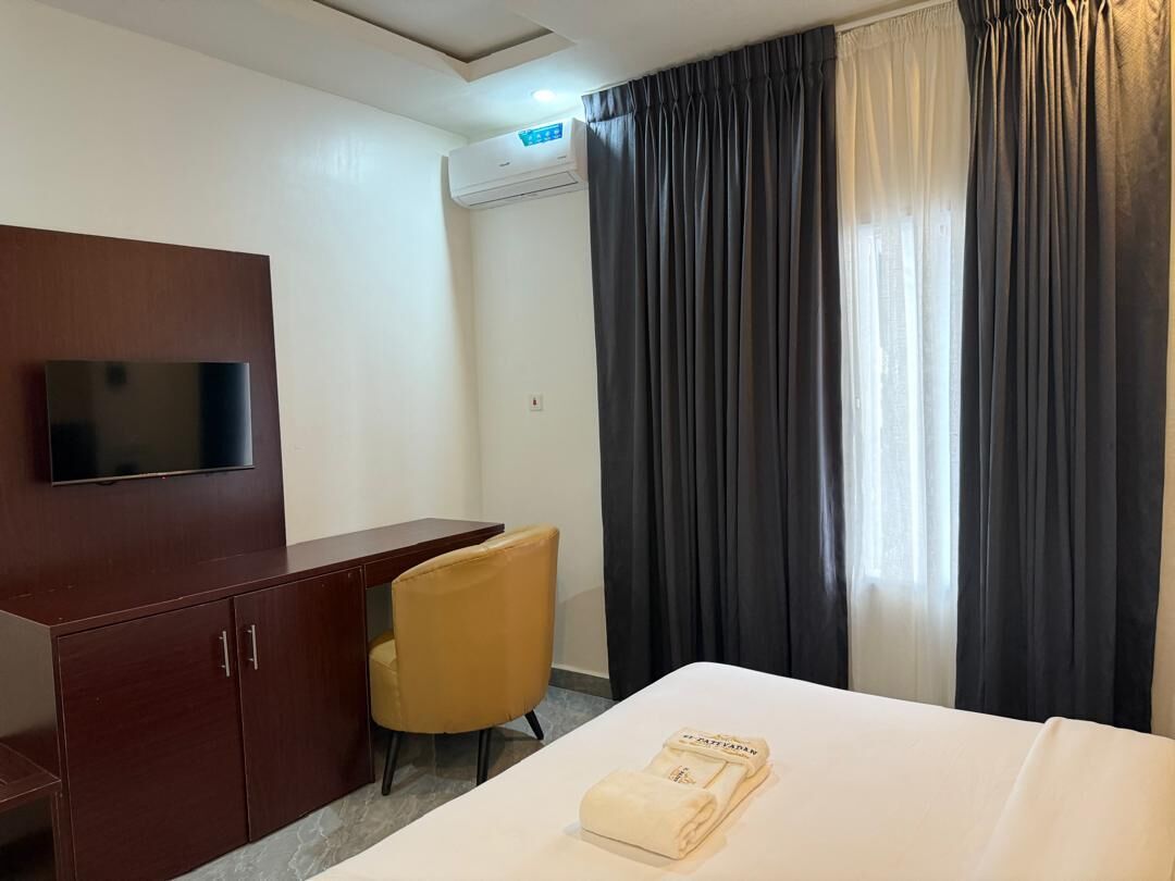 Ett hotellrum med en säng, en tv som är monterad på ett träskåp, ett skrivbord med en stol och ett fönster med gardiner.