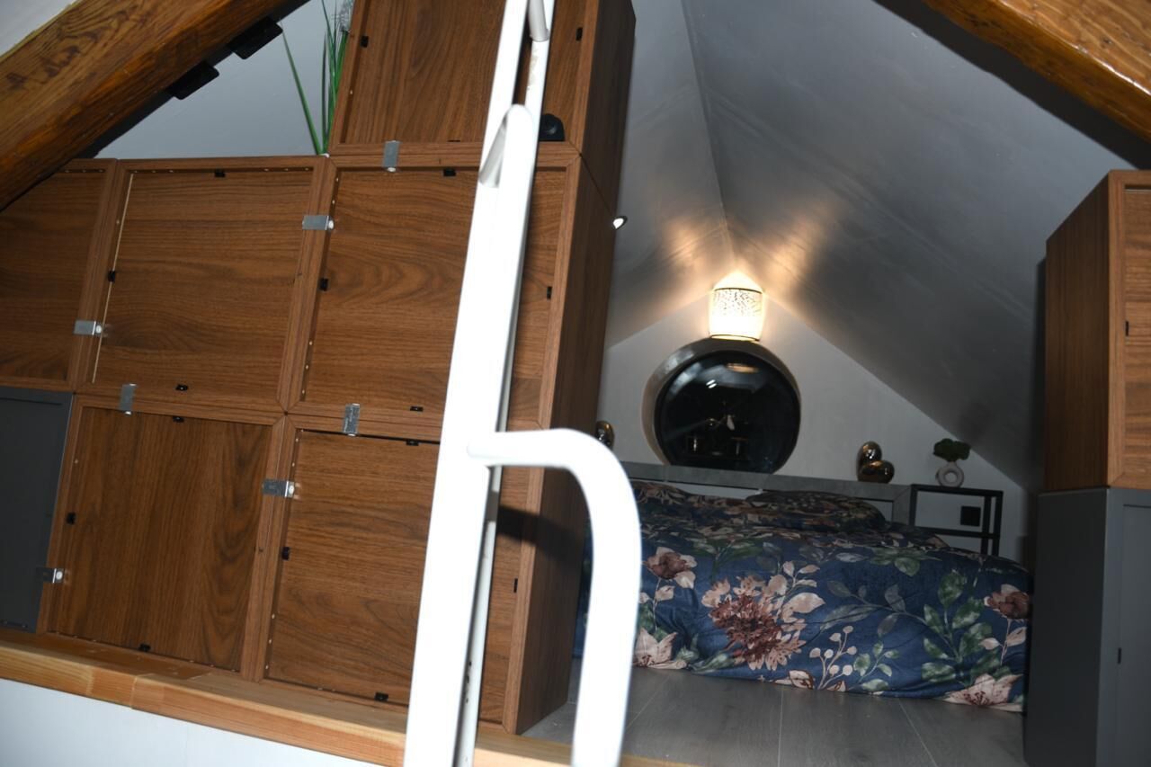 Une chambre avec un plafond en pente, des armoires en bois, un lit recouvert d’une couvre-lit à motifs floraux et un luminaire fixé au mur.