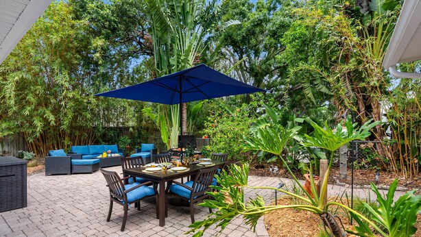 An outdoor patio with a table and chairs, surrounded by lush greenery and a blue umbrella. - Bay to Bay Bungalow | Sleeps 6 (Tampa)