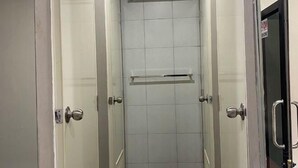 A narrow, tiled shower stall with a horizontal shelf and a door with a handle. - Hide Hostel (Koh Phangan)