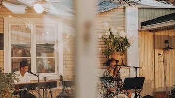 Una actuación musical al aire libre con una banda, un set de batería y un teclado, con un edificio de madera con porche como fondo.