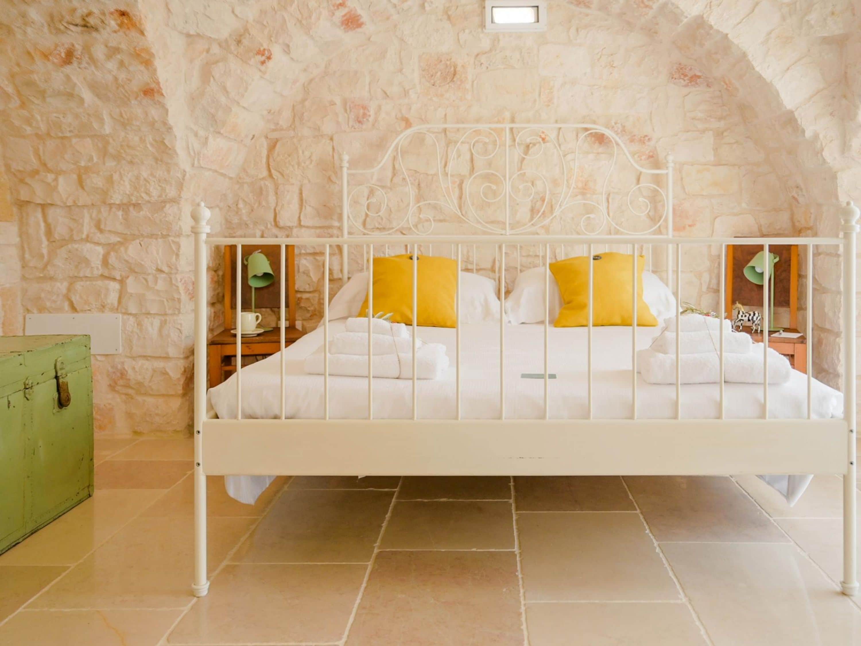A bed with white bedding and yellow pillows in a room with stone walls and a tiled floor.