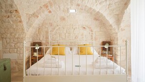 A bed with white metal headboard and white bedding, set against a stone wall with an arched ceiling.
