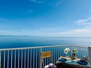 Un balcone con vista sul mare, una sedia di legno, un tavolo apparecchiato con un vaso di fiori e un cappello.