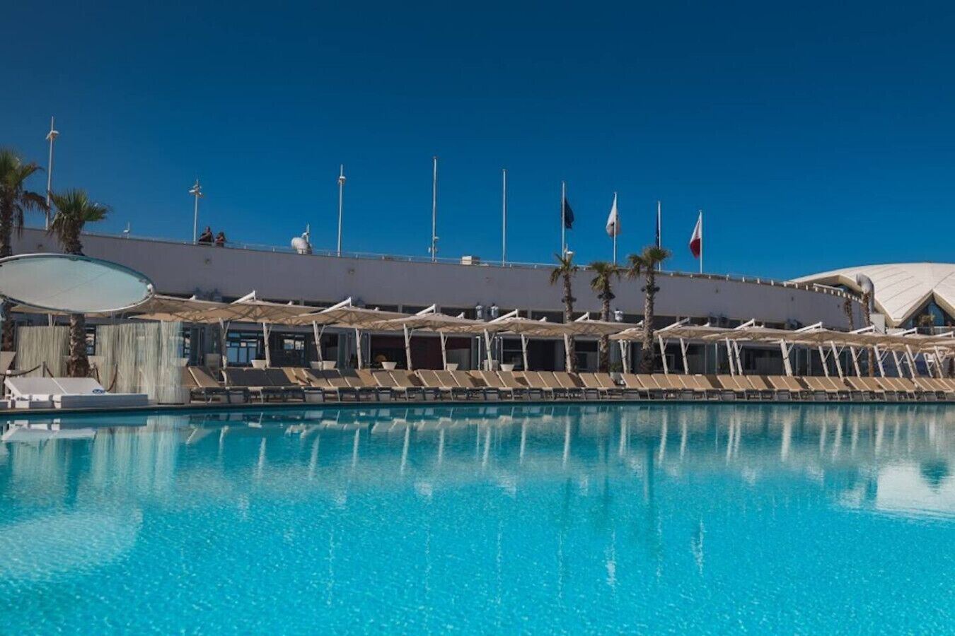 Una gran piscina con sillones y sombrillas, rodeada por un edificio con banderas y palmeras.