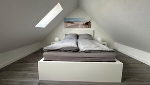 A bedroom with a sloped ceiling, a bed with gray bedding, two bedside tables with lamps, and a beach scene painting on the wall.