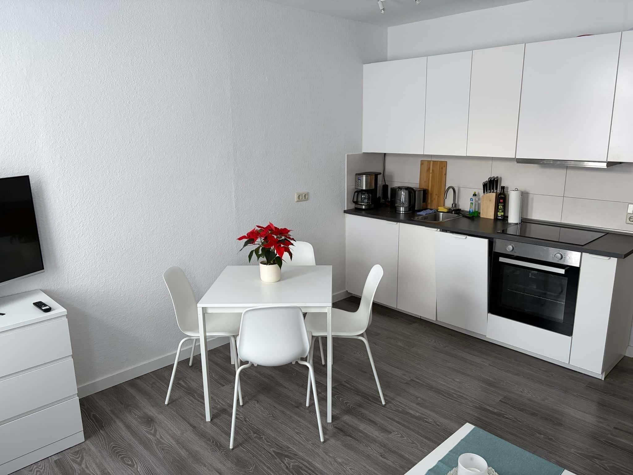 A modern kitchen with white cabinets, a dining table with chairs, a coffee maker, and a TV on a stand.