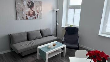 Una sala moderna con un sofá gris, una mesita de centro blanca, una lámpara de pie y una ventana con persianas.