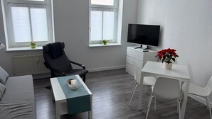 A modern living room with a sofa, a TV, a dining table, and two windows with blinds. - Close to downtown | Parking | Up to 4 guests (Chemnitz)