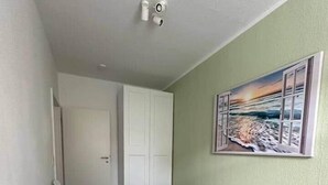A bedroom with a bed, a white chair, a door, and a painting of a beach scene. - Close to downtown | Parking | Up to 4 guests (Chemnitz)