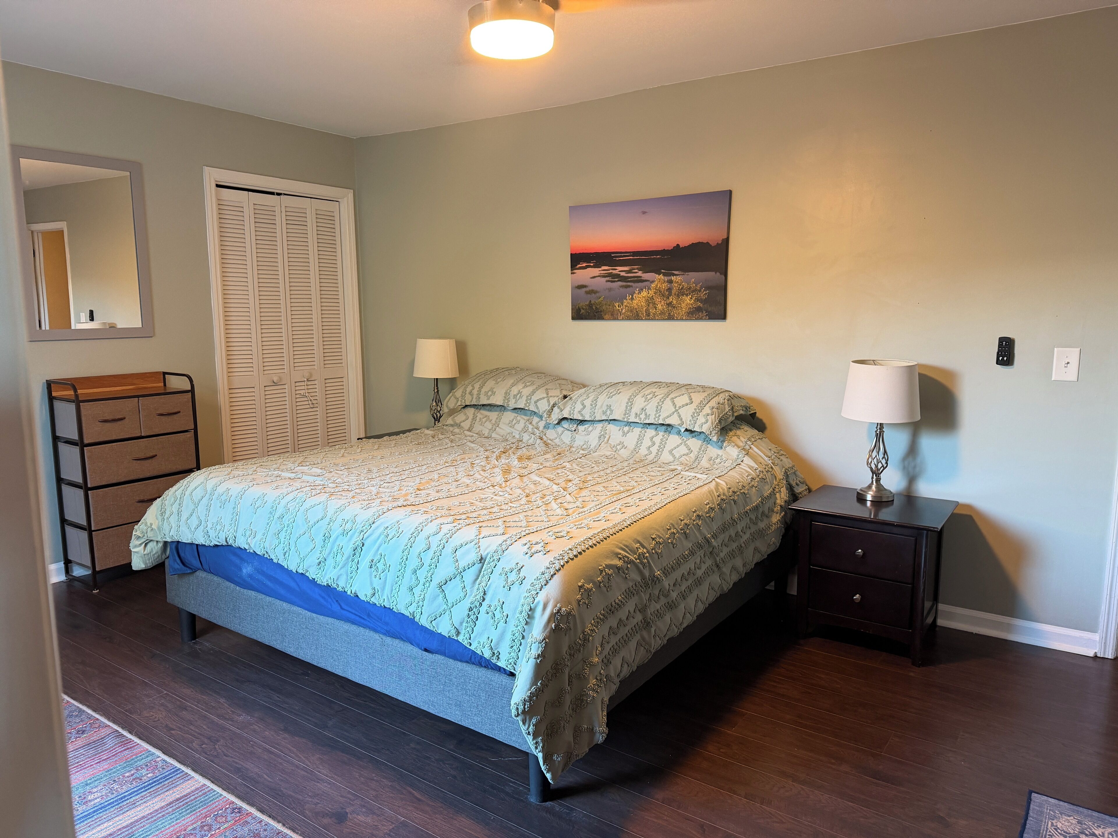 Een slaapkamer met een bed, een dressoir, nachtkastje, lamp en een schilderij aan de muur.