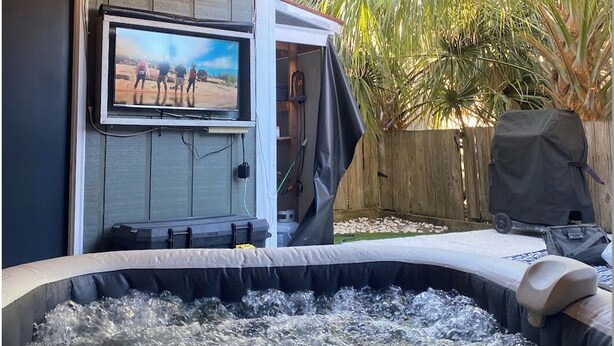 A hot tub with a television mounted on a wooden wall, surrounded by a wooden deck and a garden with palm trees. - Luxury Townhouse near Downtown and Beaches with Hot Tub (Mount Pleasant)