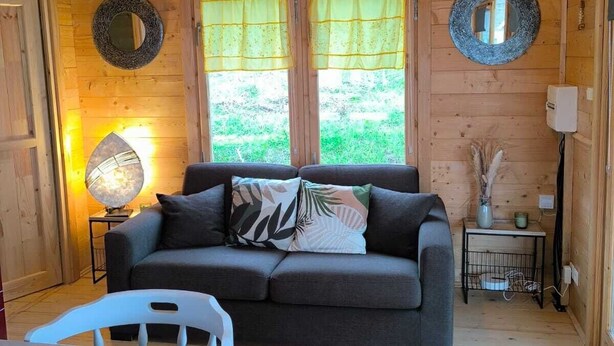 Un accogliente interno di una baita con soffitto in legno, un divano con cuscini, un tavolo con sedie e due finestre con tende.
