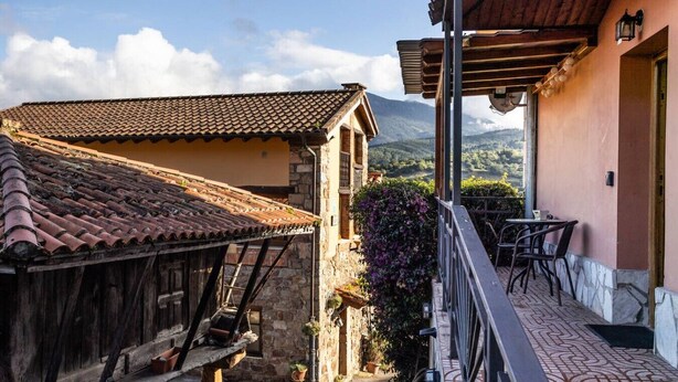 A traditional building with a tiled roof, a balcony with outdoor seating, and a view of mountains in the background. - Peaceful Village Escape (Sales)