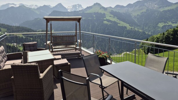 Un balcon agrémenté de meubles en osier, d’une balançoire en bois et d’une table, offrant une vue sur un paysage montagneux.