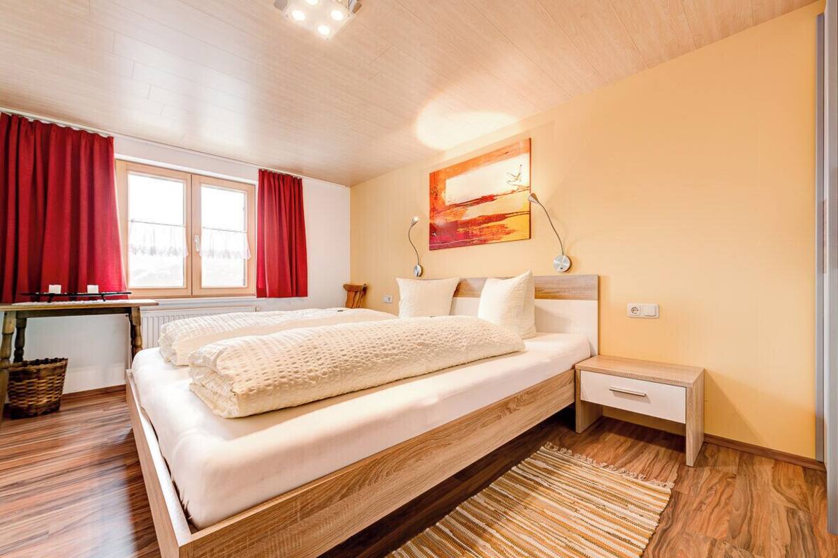 A bedroom with a wooden bed, a nightstand, a window with red curtains, and a painting on the wall.