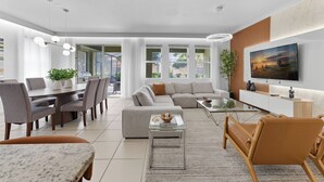 A modern living room with a dining area, a sofa set, a TV mounted on the wall, and a large window with a view of the outdoors. - 7049 Solterra 6 bedrs 18guests pool summer kitchen (Davenport)