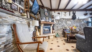 A cozy living room with a stone fireplace, a striped armchair, and a bookshelf filled with books and decorative items. - The Village Retreat - Al Llagar (Sales)