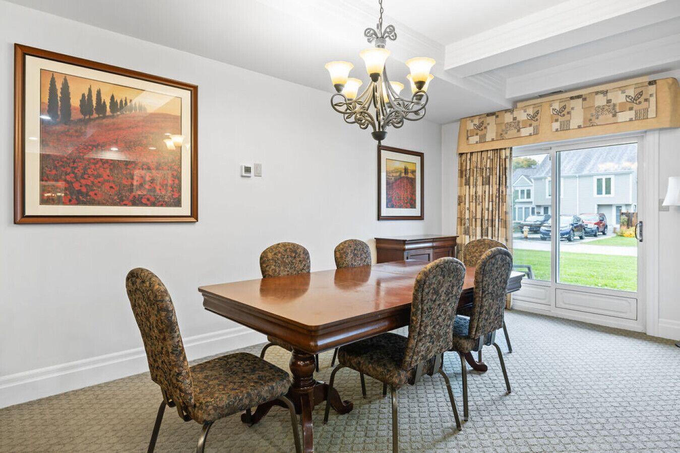 A dining room with a wooden table and patterned chairs, a chandelier, and two framed artworks on the walls.