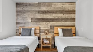 A bedroom with two beds, wooden headboards, a small bedside table with a lamp, and a wall-mounted shelf.
